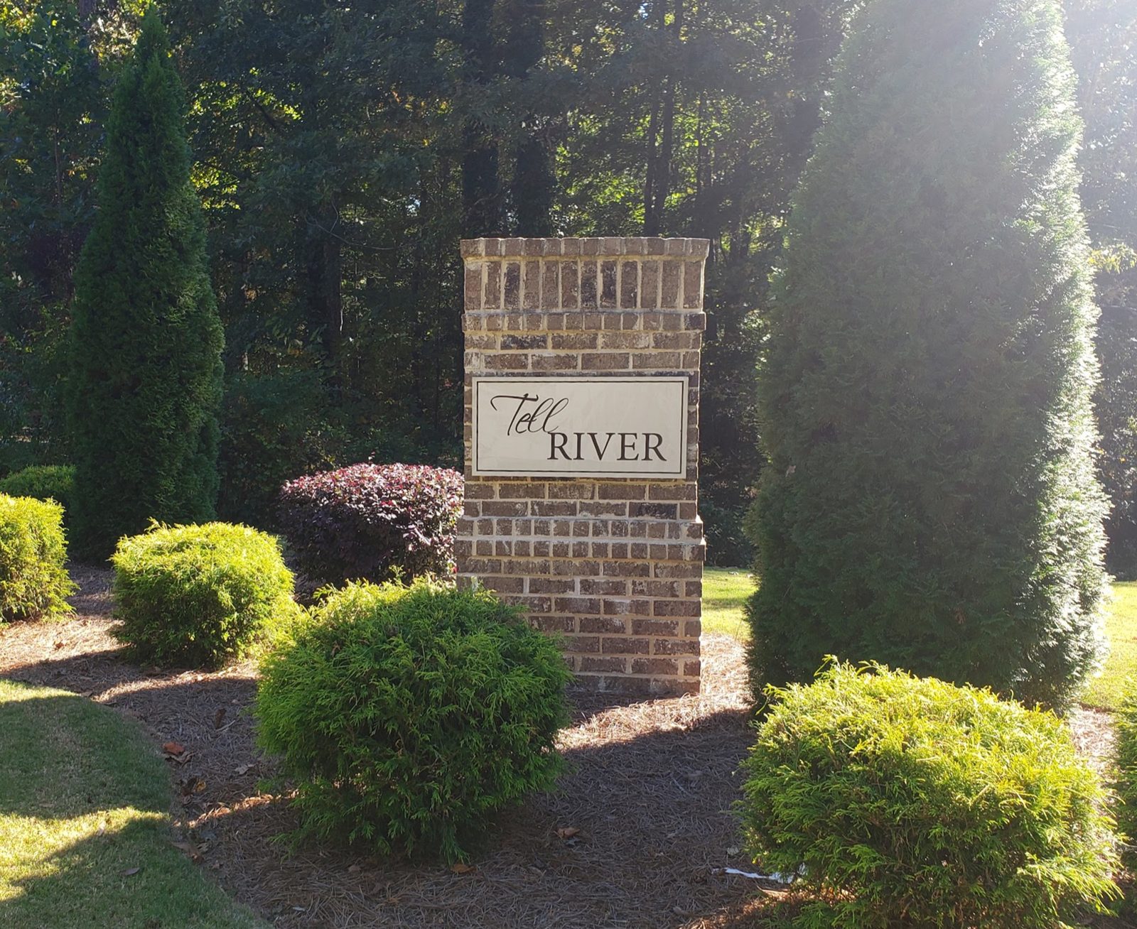 Brick entrance monument with a Tell River sign, surrounded by shrubs and evergreens